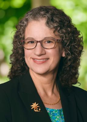 Headshot of white woman with curly brown hair and brown rimmed glasses wearing a black blazer