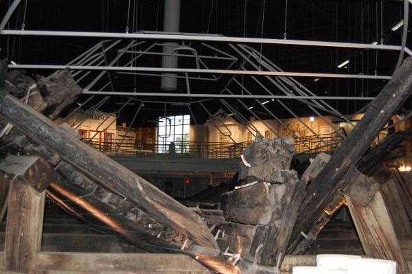 Interior view of the CSS Neuse Museum showing the preserved remains of the CSS Neuse ironclad. Large, weathered wooden timbers from the ship's hull fill the foreground, with the skeletal frame of the vessel extending toward the back of the gallery. Overhead lighting illuminates the exhibit, highlighting the size and construction of the Civil War ironclad. An elevated walkway and interpretive displays are visible in the background, allowing visitors to view the ship from multiple perspectives.