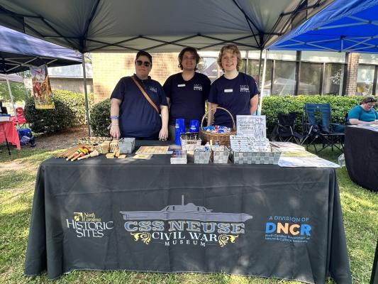 Three people in blue shirts stand behind a table displaying historical information and artifacts, with tents and other vendors in the background.