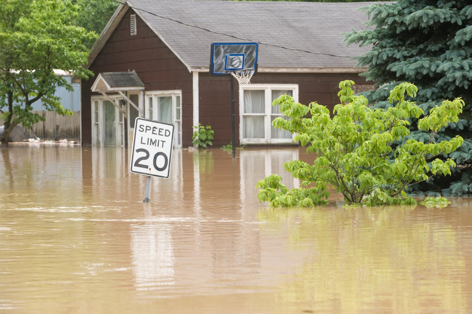 Floodwater surrounds a brick home on a residential street, partially submerging a nearby speed limit sign