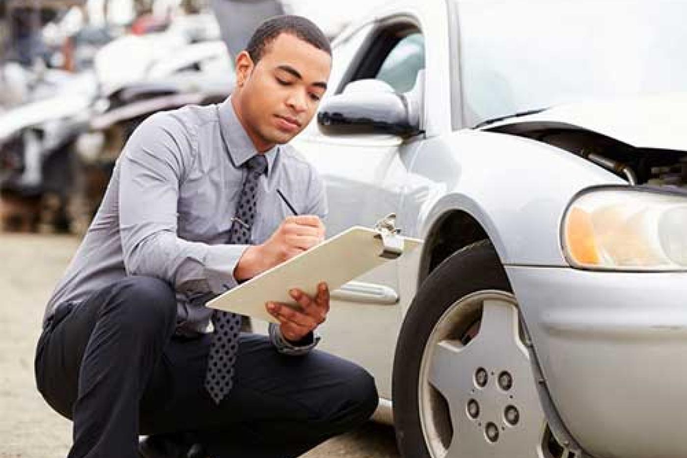 Man kneeling beside crashed car taking notes on a clipboard.