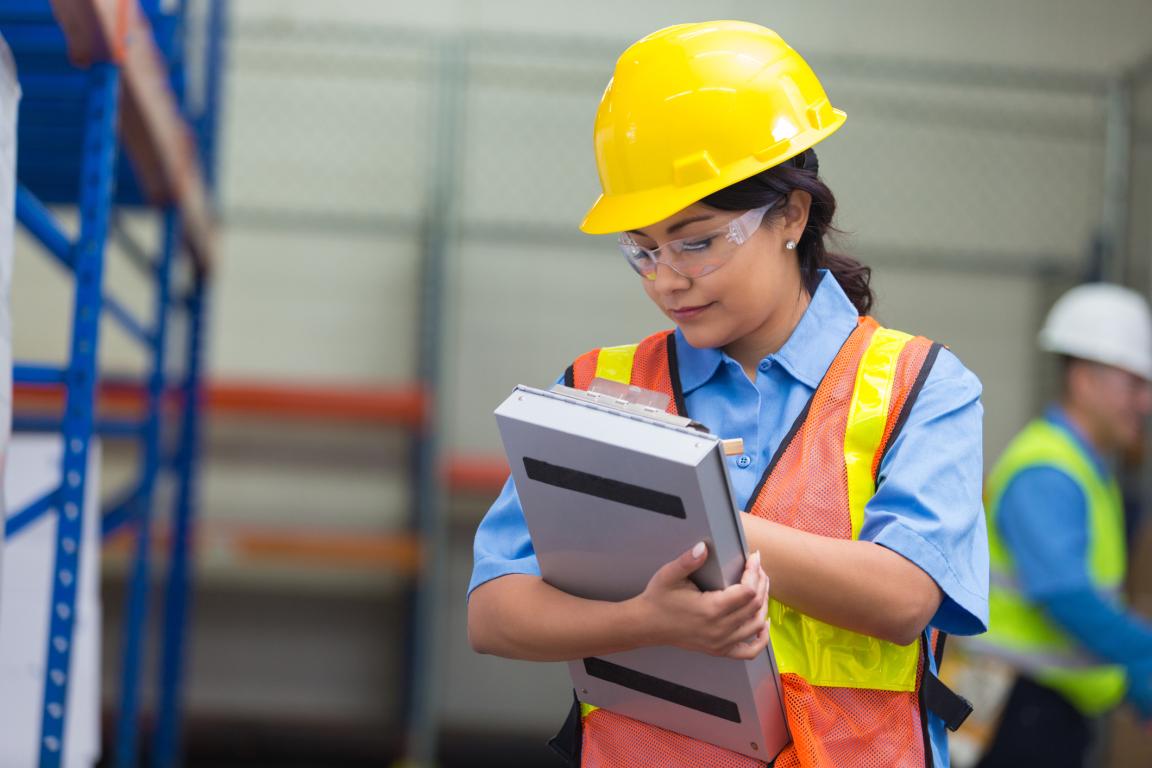 A construction worker writes on a clipboard while wearing a hardhat, safety vest and safety glasses