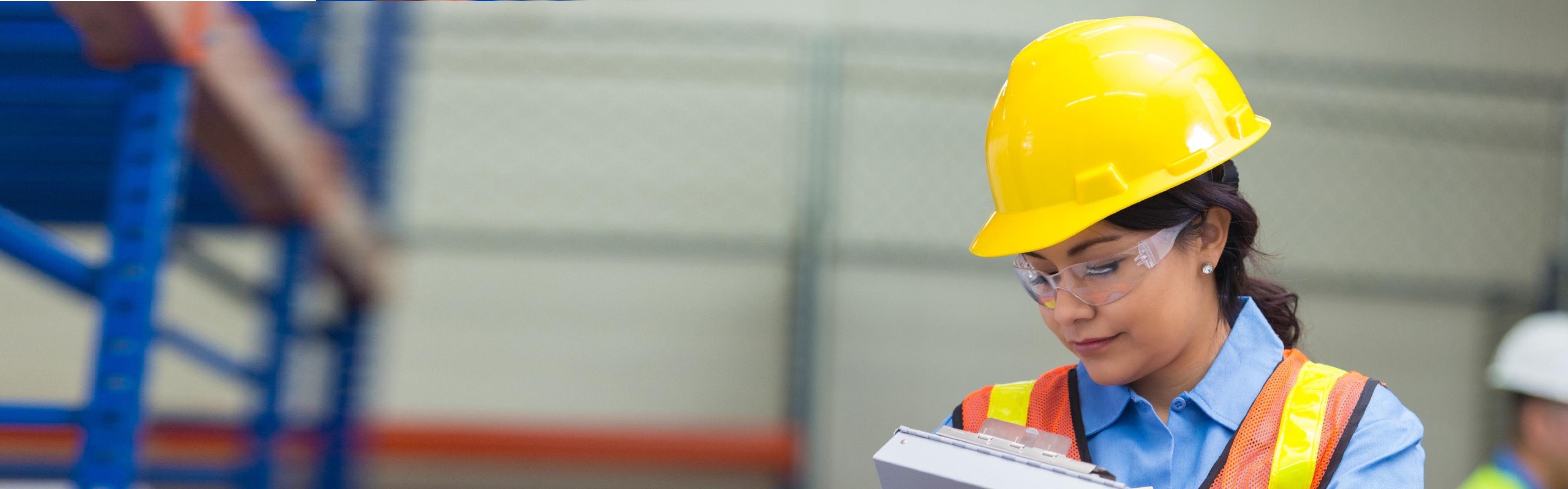 Employee wears a hard had and protective glasses, marks a clipboard next to a conspicuous scaffold structure