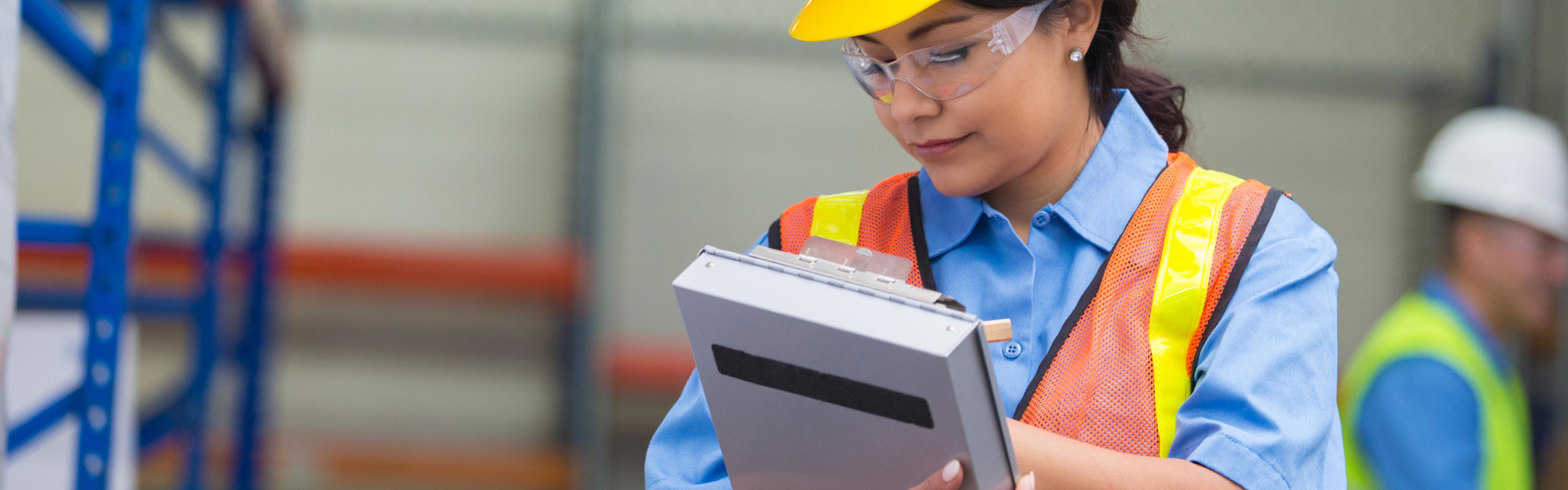 A construction worker writes on a clipboard while wearing a hardhat, safety vest and safety glasses
