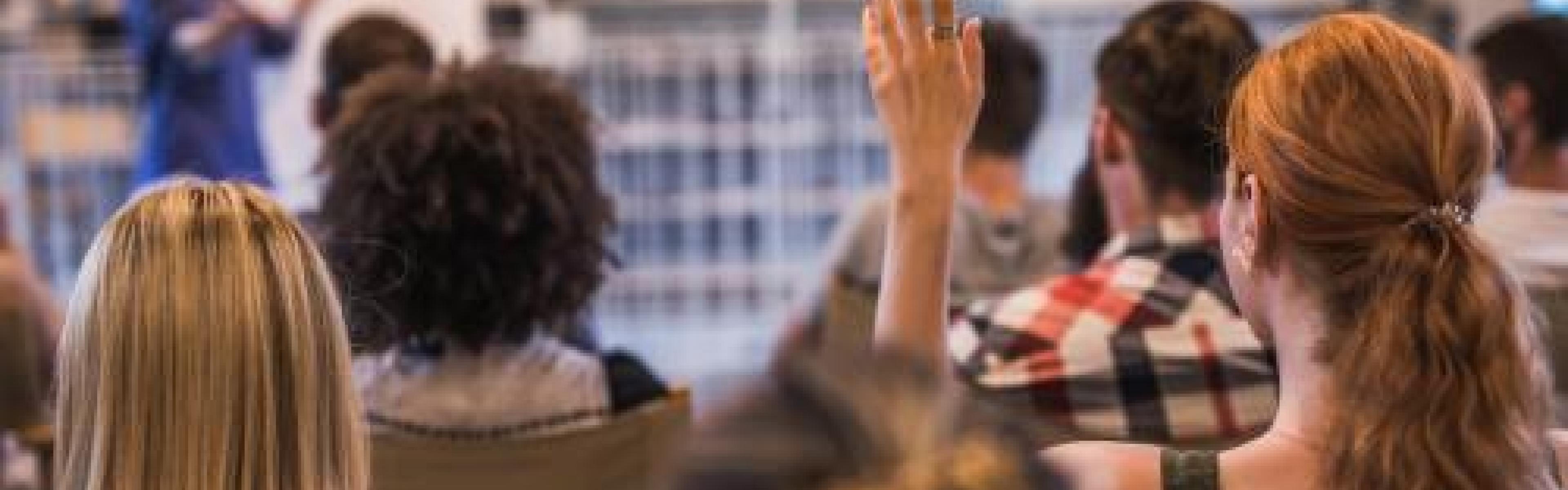 A classroom full of people watching a presentation and one person is raising their hand