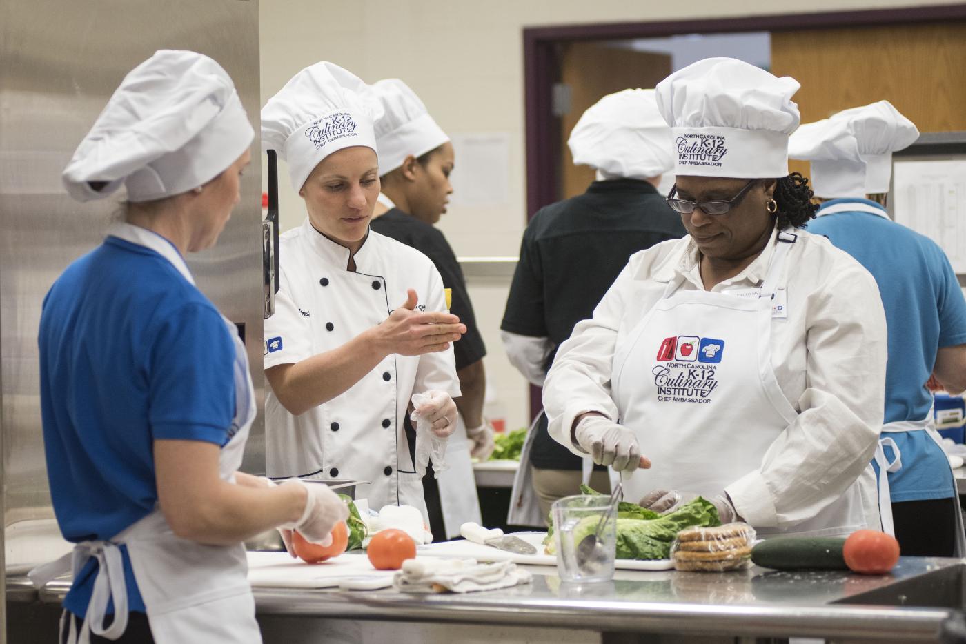 Two school nutrition professionals chopping vegetables on a work table while another coaches them.