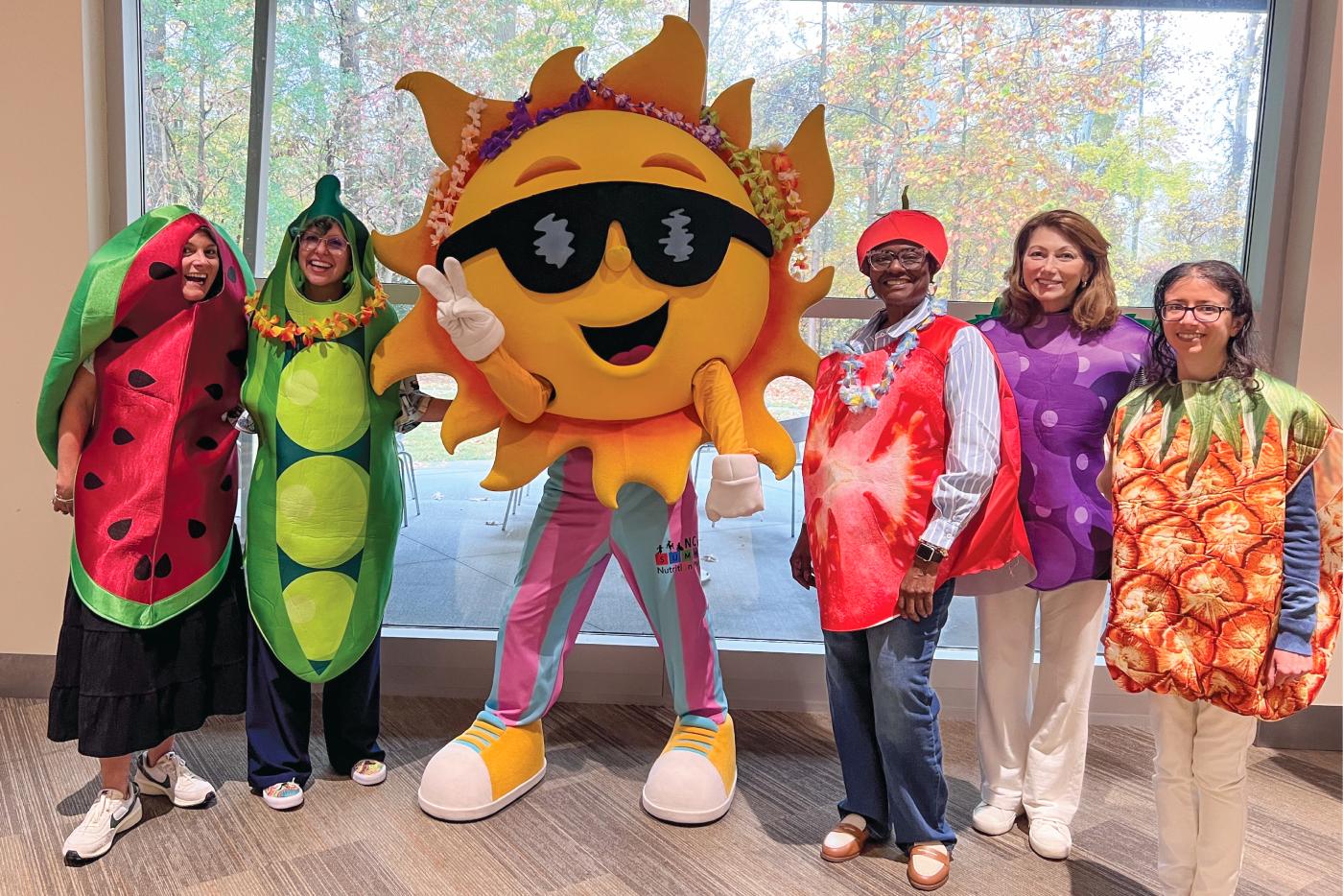 School nutrition staff dressed as fruits and vegetables surround a person wearing a sun costume.