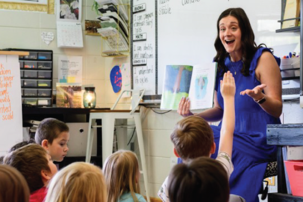A teacher sitting in a chair reads a book to her students, who are sitting on the floor.