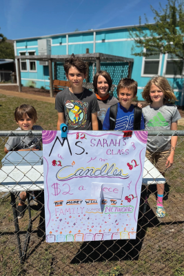 Island Montessori students pose behind a sign that says “Ms. Sarah’s Class. Candles - $2 apiece. The money will go to Family Support Networks.”
