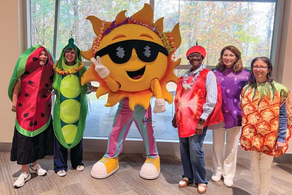 School nutrition staff dressed as fruits and vegetables surround a person wearing a sun costume.