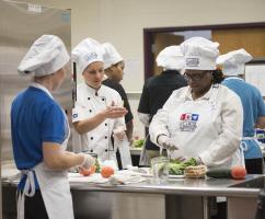Two school nutrition professionals chopping vegetables on a work table while another coaches them.