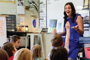 A teacher sitting in a chair reads a book to her students, who are sitting on the floor.