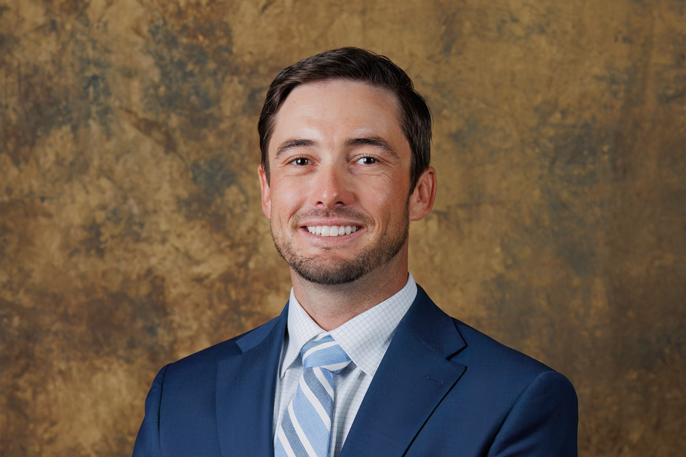 justin boyd poses in a blue suit and tie in front of a mottled bronze background