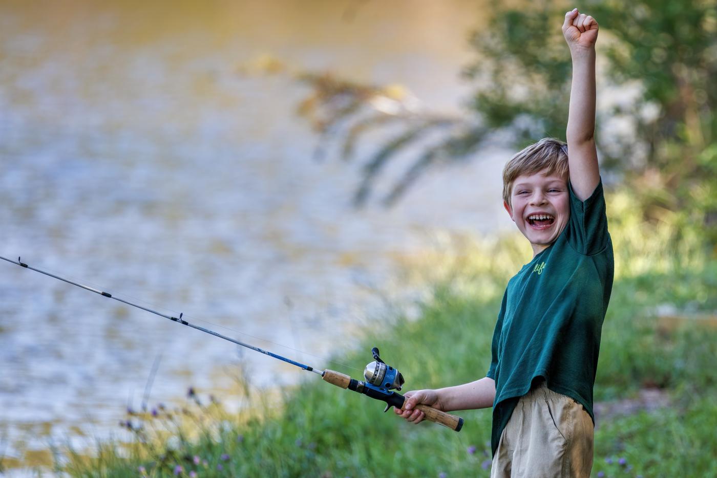 a young boy holds a fishing rod with one hand and raises his other arm over his head with a joyous expression