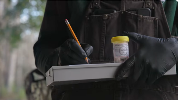A person is seen holding a clip board and writing on it with a pencil while holding a CWD specimen container.