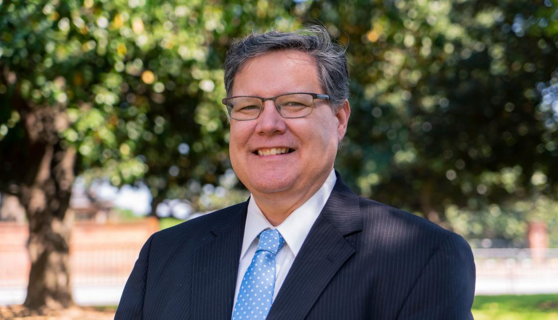 Bill Holmes Image of a man in a blue suit standing outside in front of two trees.