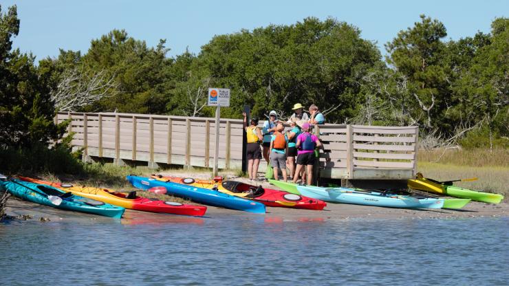 People stand on the end of a boardwalk next to kayaks along the shoreline.