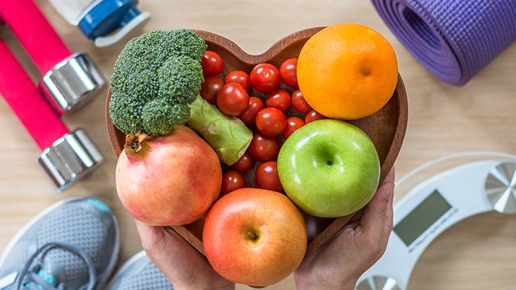 Hands holding a bowl of fruit surrounded by water bottle and exercise equipment