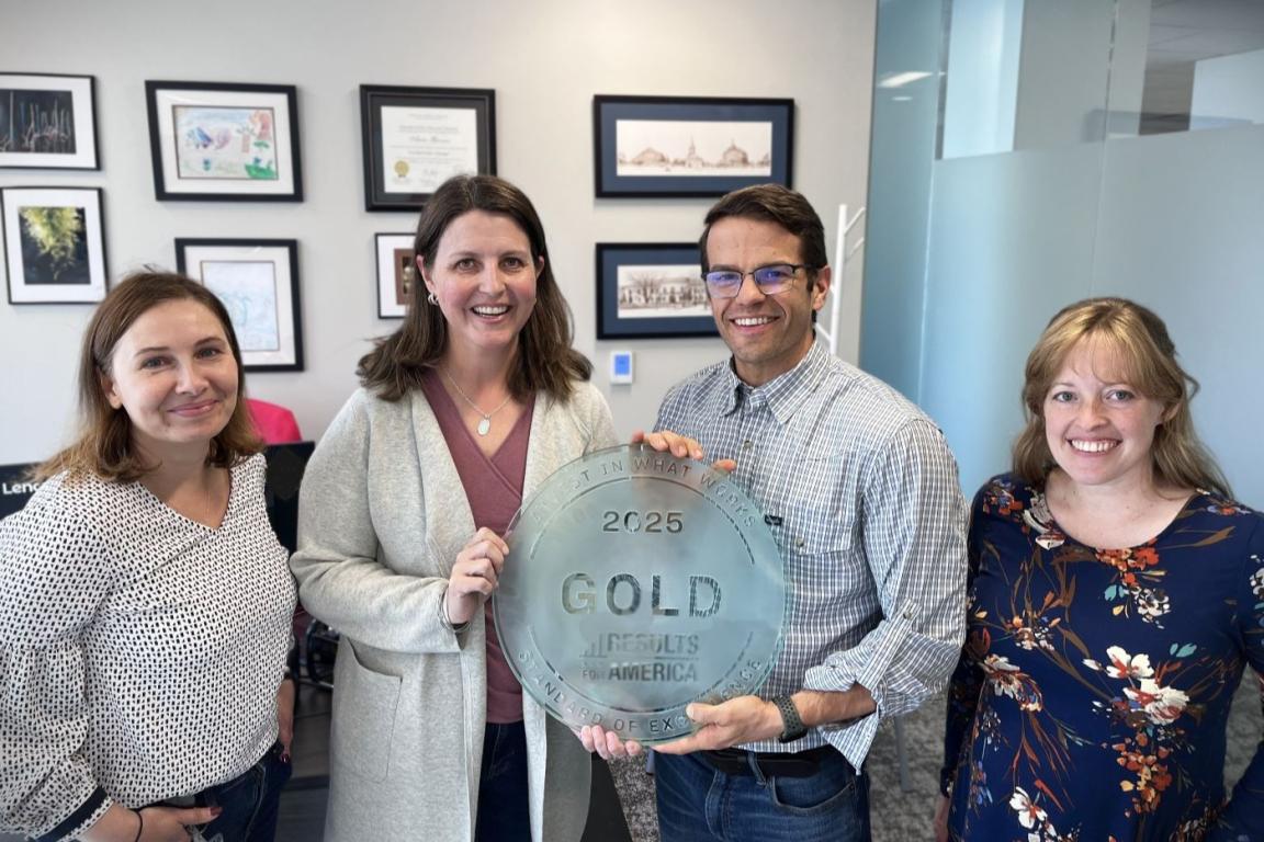 Three women and a man stand shoulder to shoulder, smiling. Two people in the center hold a circular plaque