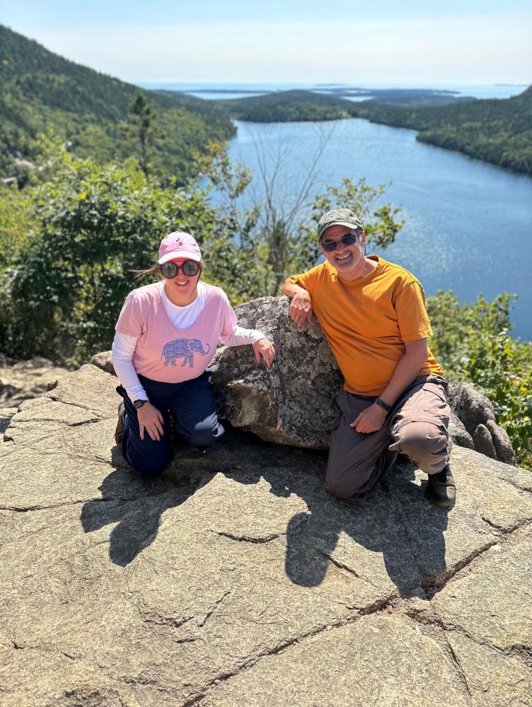 Woman and man in hats and sunglasses smiling, kneeling on a ledge overlooking lake between rolling green hills