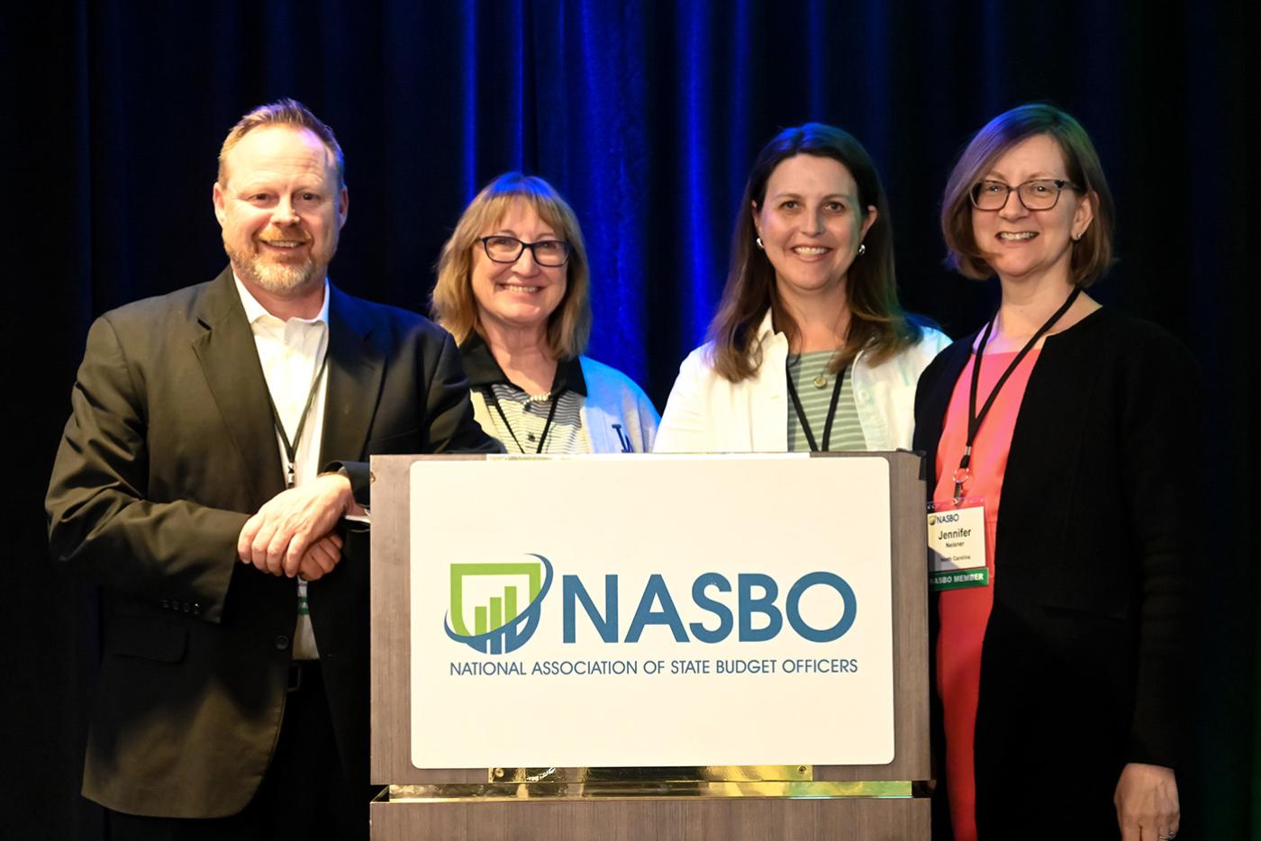 Four people behind a podium smiling. Podium sign says NASBO