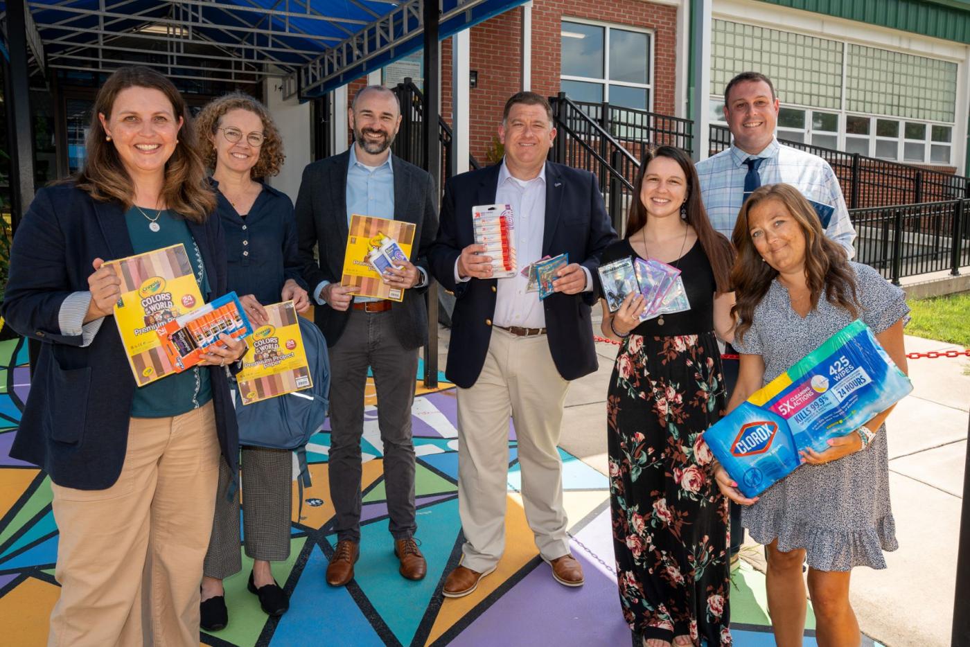 Group of people standing in front of entrance of a school smiling. Each is holding school supplies like paper, glue, wipes and earbuds