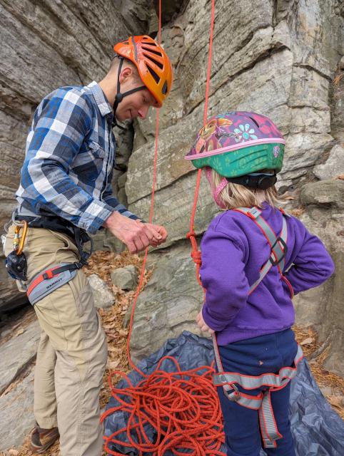 Man wearing a helmet focused on tying a rope as a little girl watches