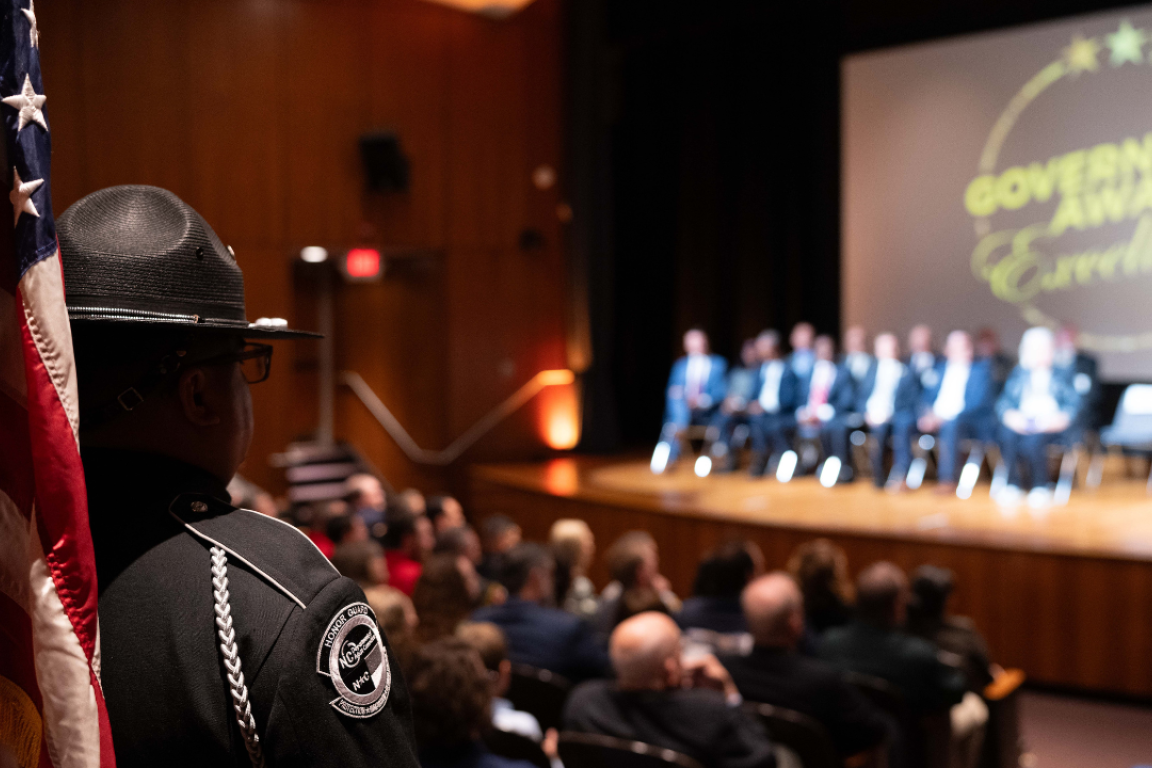 DAC Honor Guard member with American flag in foreground, stage with Governor's Awards for Excellence logo on screen in background.