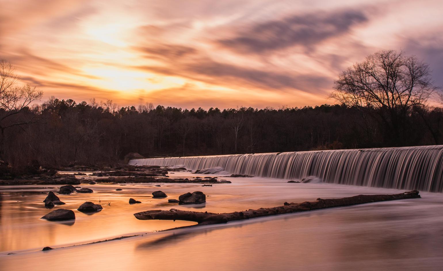 a sunset over the Bynum Dam with water spilling over into a calm river