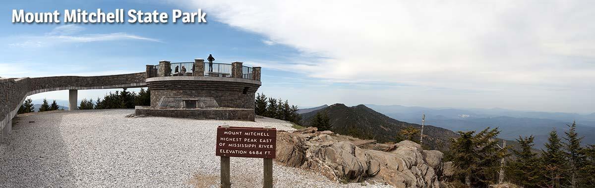Mount Mitchell State Park observation tower at the summit built of stone, with a wooden sign reading Mount Mitchell Highest Peak East of Mississippi River Elevation 6,684 Ft