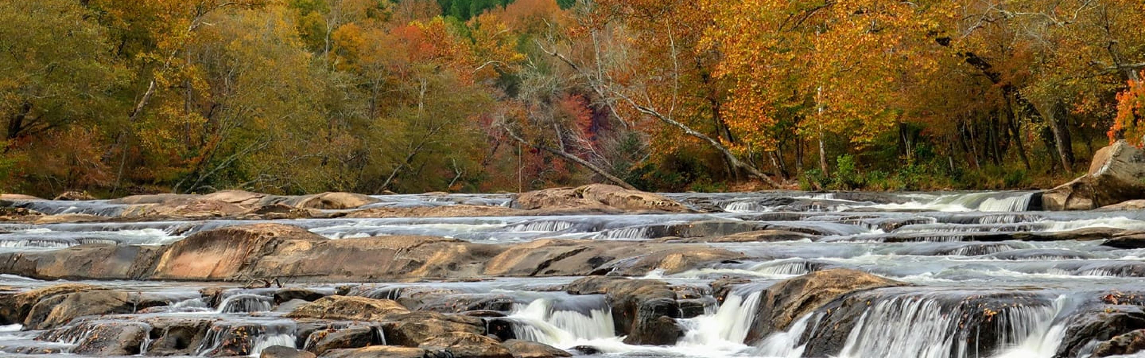 a wide river with rocks and water spilling over the rocks with fall foliage in the background