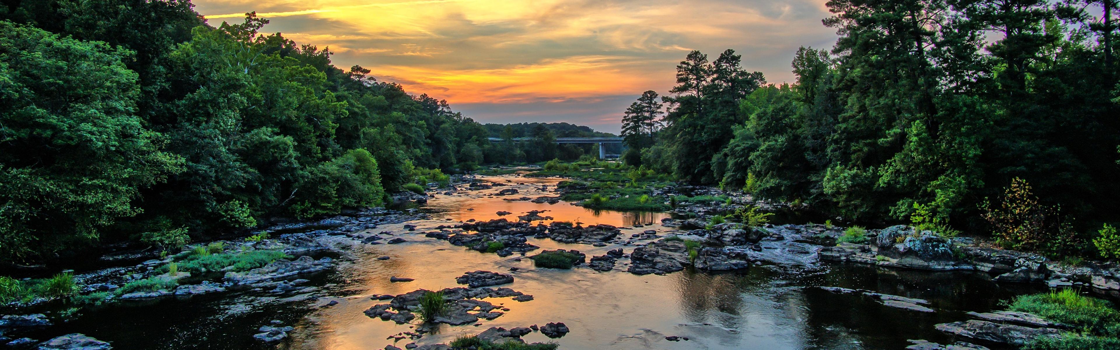 a sunset over a river being reflected back among calm water, rocks, and vegetation on both sides.