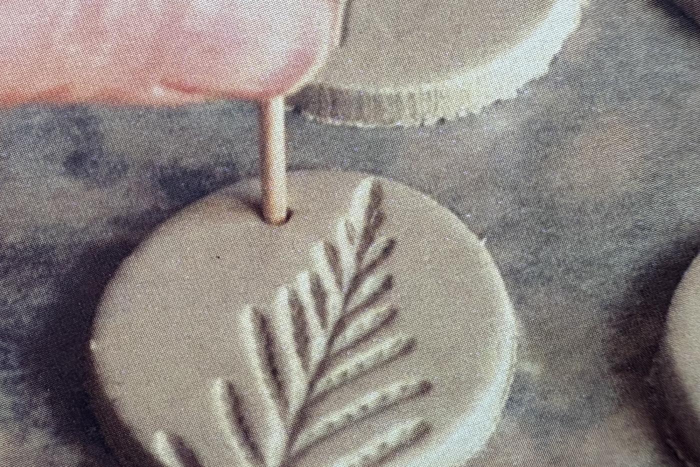 A clay ornament with the imprint of a fern is being shaped.