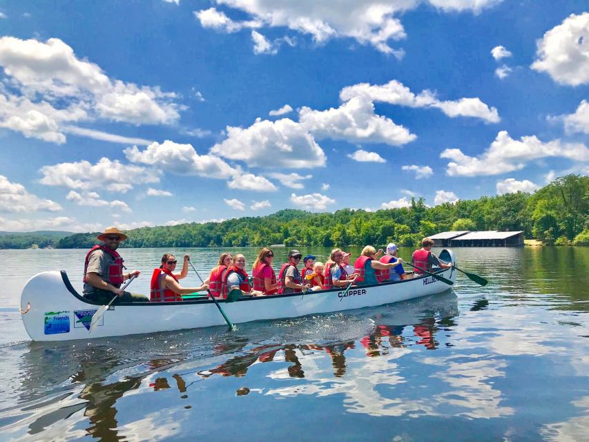 A group of students and a park ranger in a big canoe paddling across a calm lake with a blue sky and trees in the background