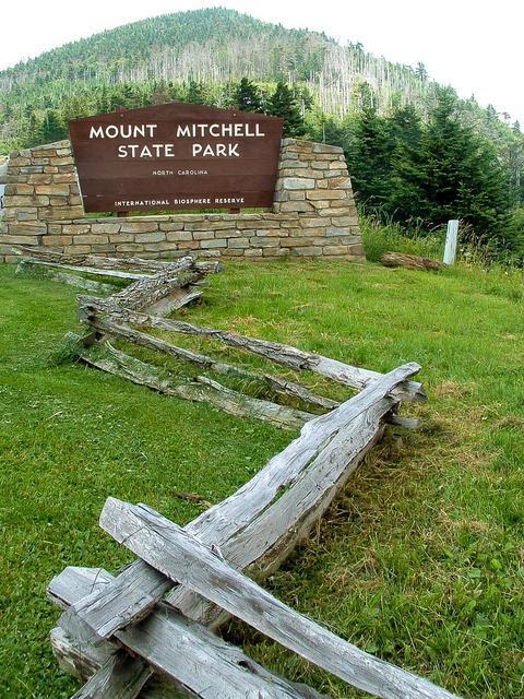 Mount Mitchell State Park entrance sign supported by stacked stone walls with a rustic split rail wooden fence in the foreground