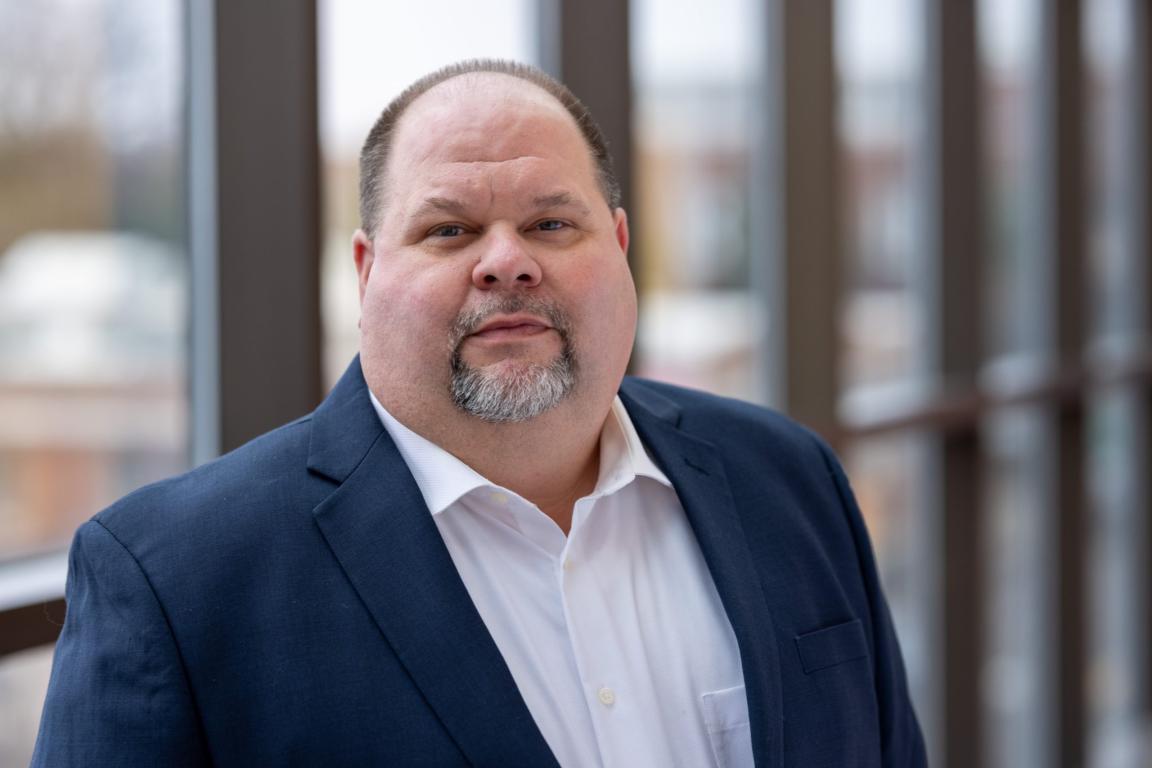 Portrait of a middle-aged man in a navy suit and white shirt, standing indoors with a blurred windowed background.