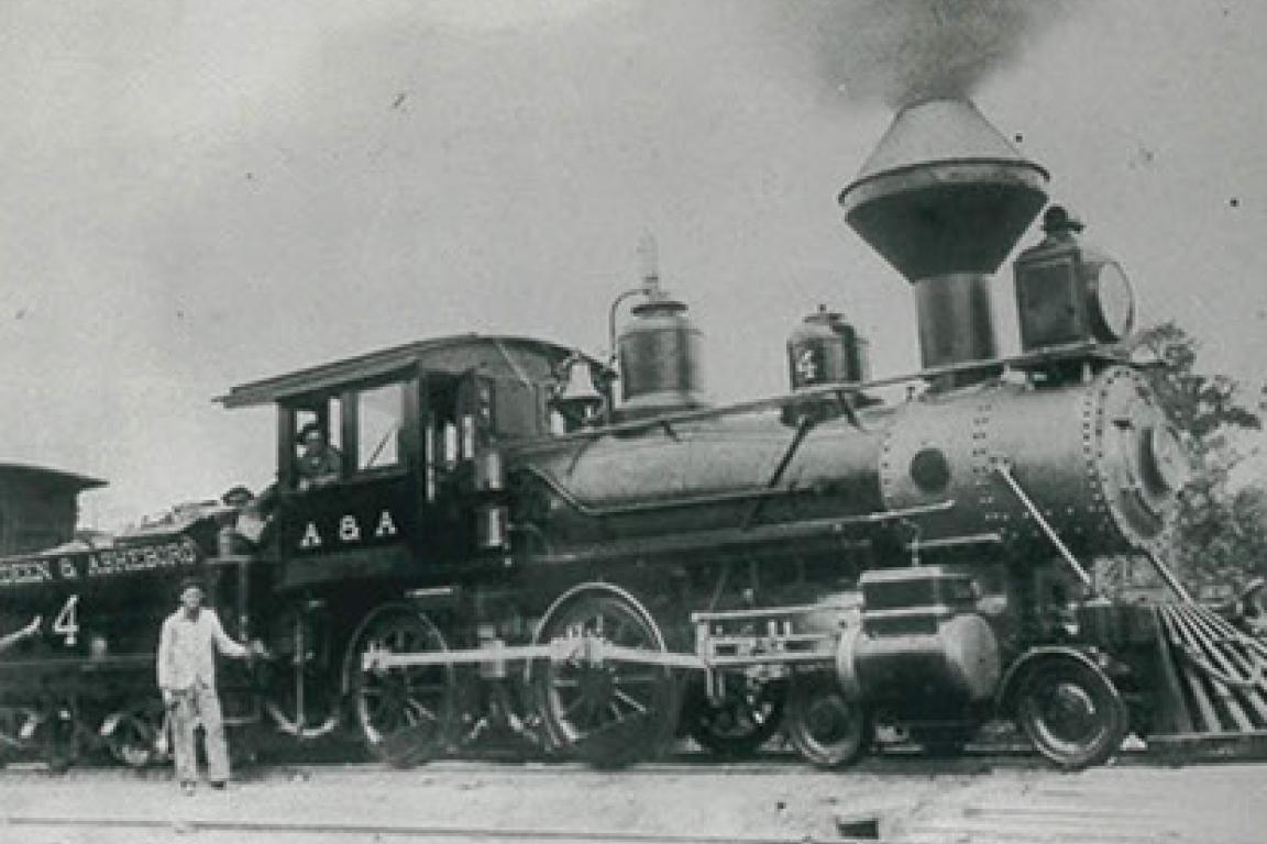 Black-and-white photo of a vintage steam train labeled “A&A,” with a man standing beside it on railroad tracks.