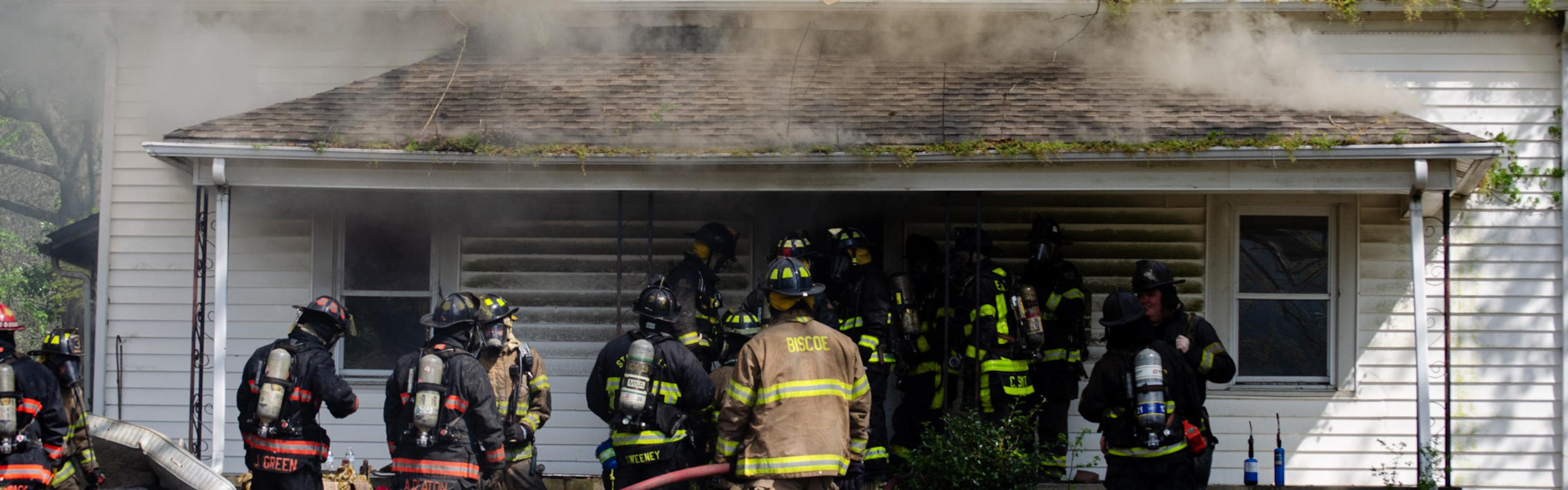 Firefighters conduct a live fire training exercise at a house, with smoke rising as crews practice response and suppression.