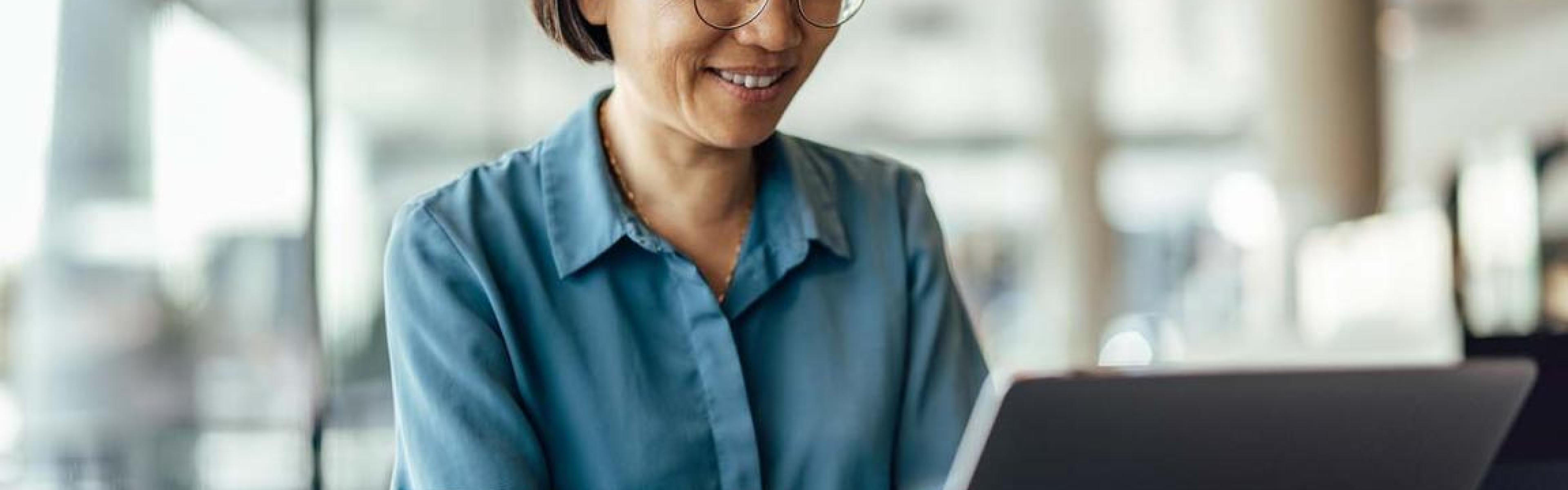 A middle aged Asian woman smiles and types on a laptop in a bright, airy office.