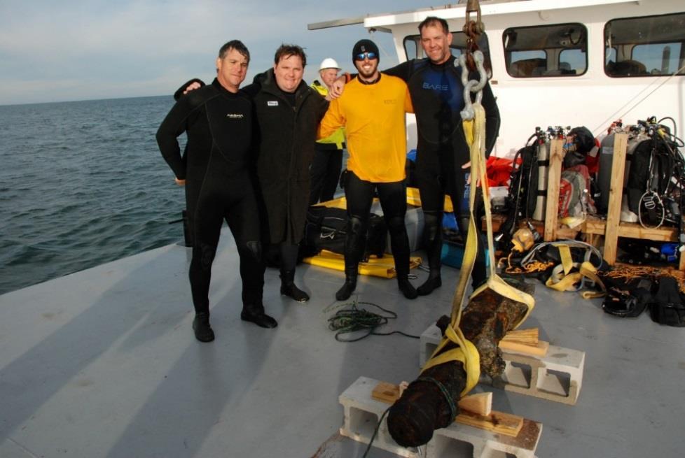 Four male team members smiling on the deck of a boat with the recovered, concreted cannon secured to wooden blocks