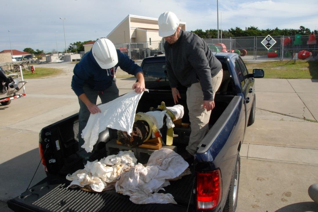 Two individuals wrapping the recovered, concreted cannon with wet rags in the back of a pickup truck