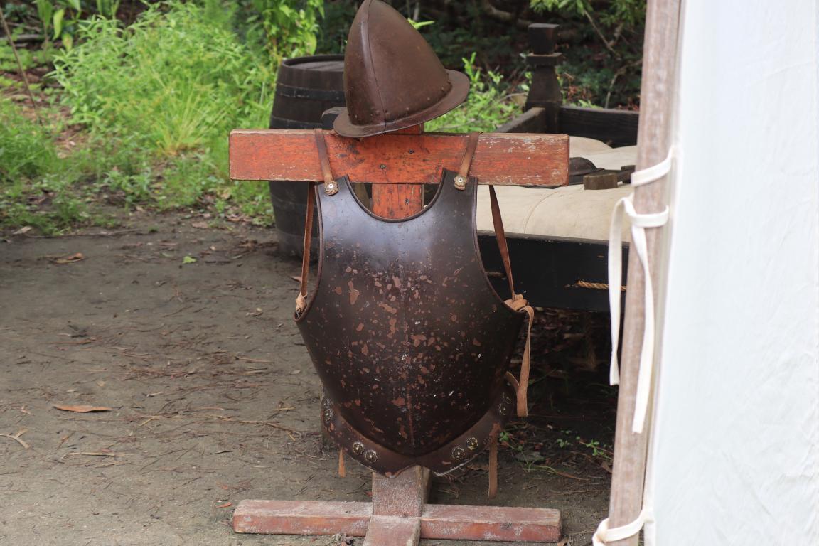 A rusted metal breastplate and helmet displayed on a wooden stand outdoors near a tent.