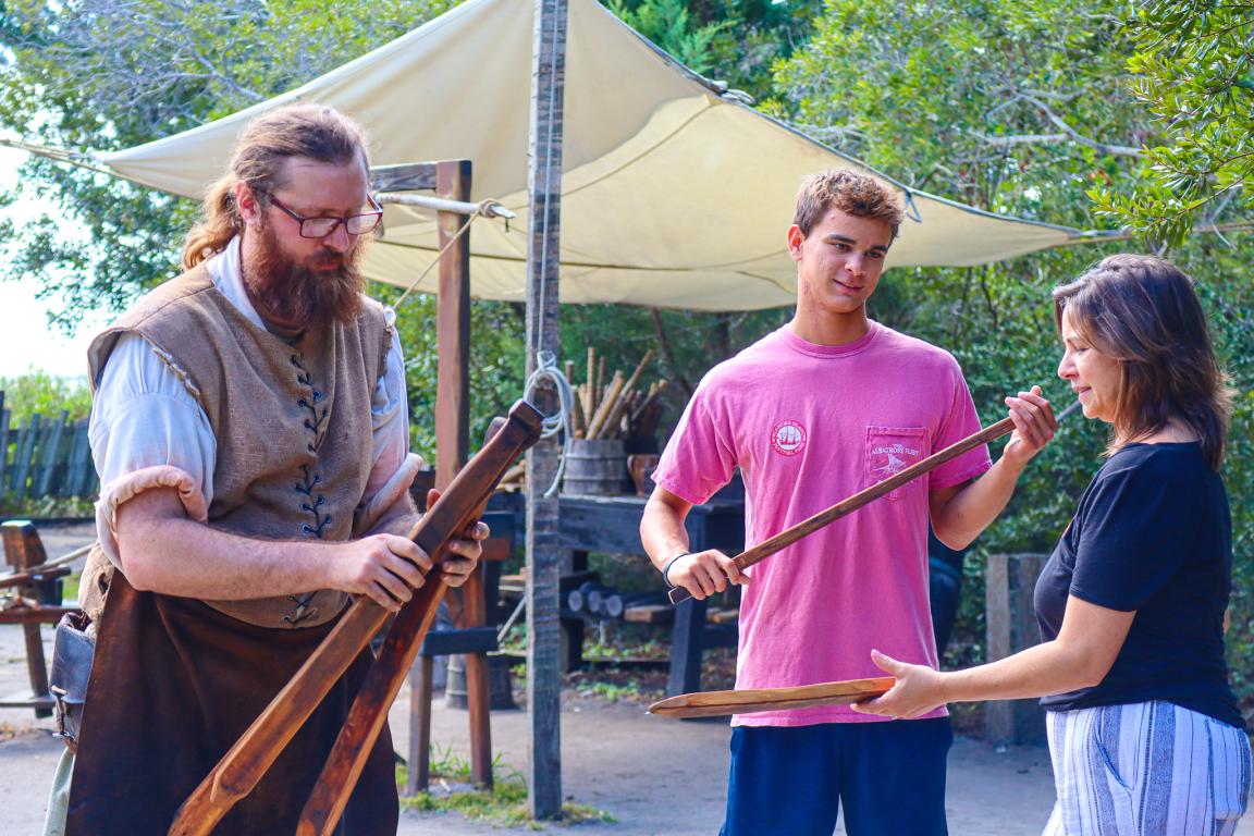 Costumed interpreter holding wooden swords and passing them out to two guest.