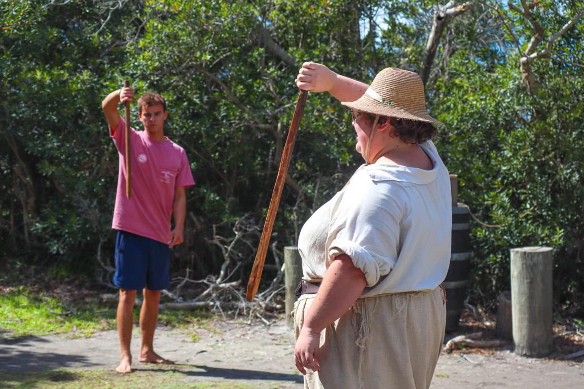 Costumed interpreter holding a wooden sword in front of their face, with a civilian in front of them doing the same motion with their wooden sword.