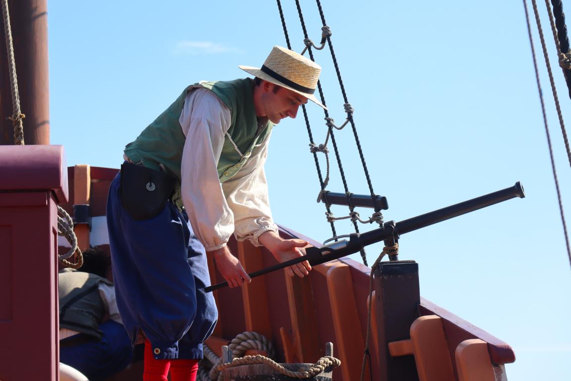 A person in historical attire, including a straw hat and vest, peers through a long telescope on a wooden ship deck against a clear blue sky.