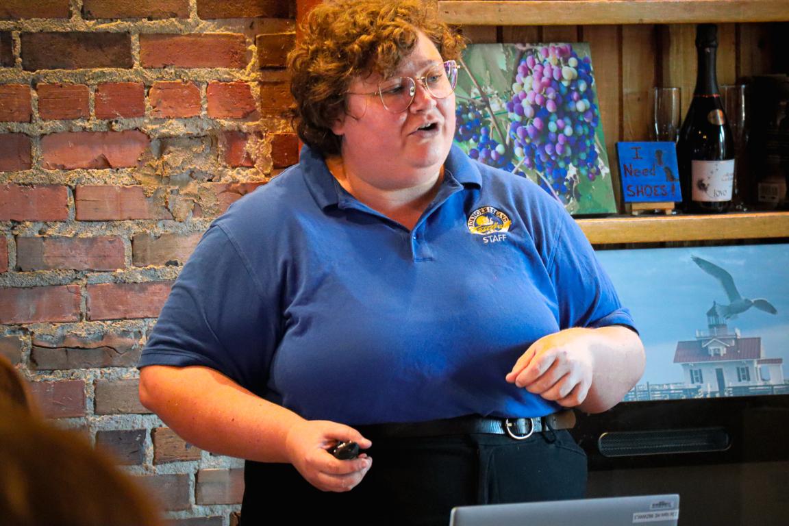  A person with curly hair and glasses speaks passionately indoors. They wear a blue staff polo shirt. The brick wall and colorful art create a warm atmosphere.