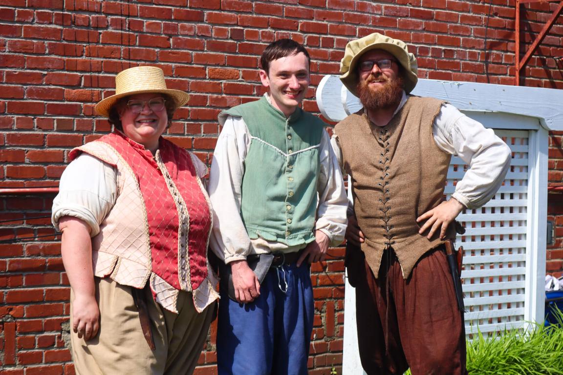 Three people dressed in historical costumes stand smiling in front of a brick wall. Their outfits are colorful, with vests and hats, conveying a cheerful, festive atmosphere.