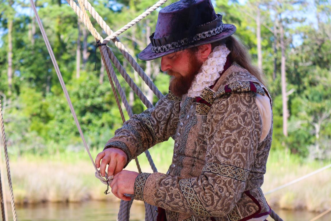 Bearded historical reenactor in an ornate Renaissance costume and tall hat ties ropes beside a wooded pond, creating a theatrical atmosphere.