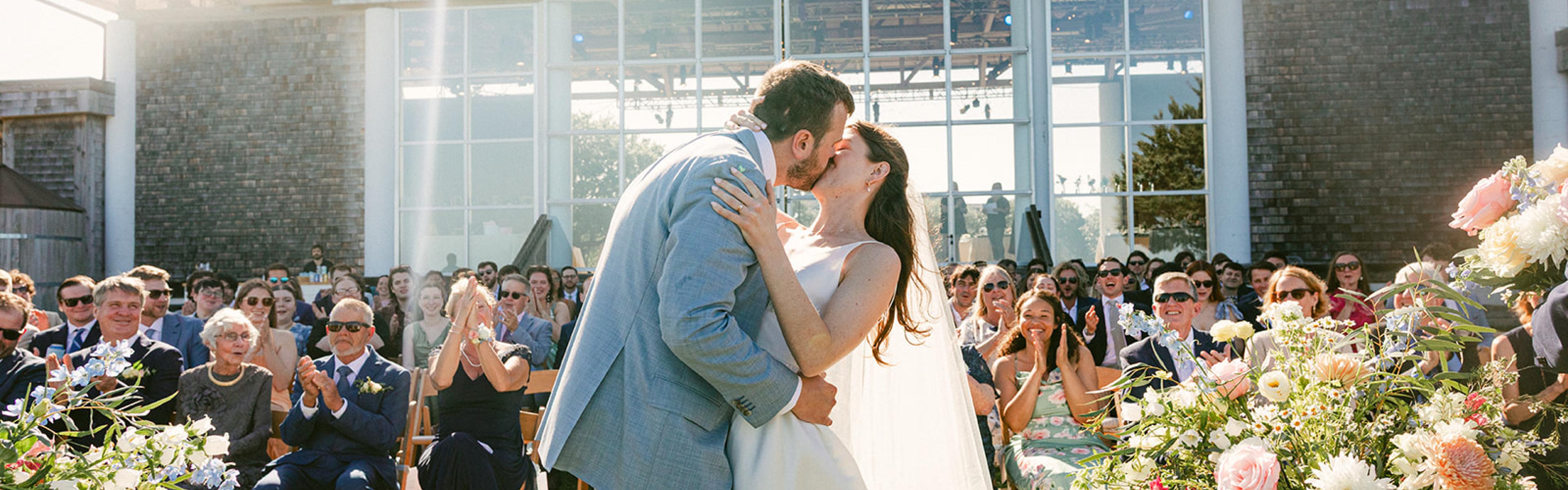 Wedding Photo: Bride and Groom kissing in front of a wooden pavilion with people in chairs clapping in the background.