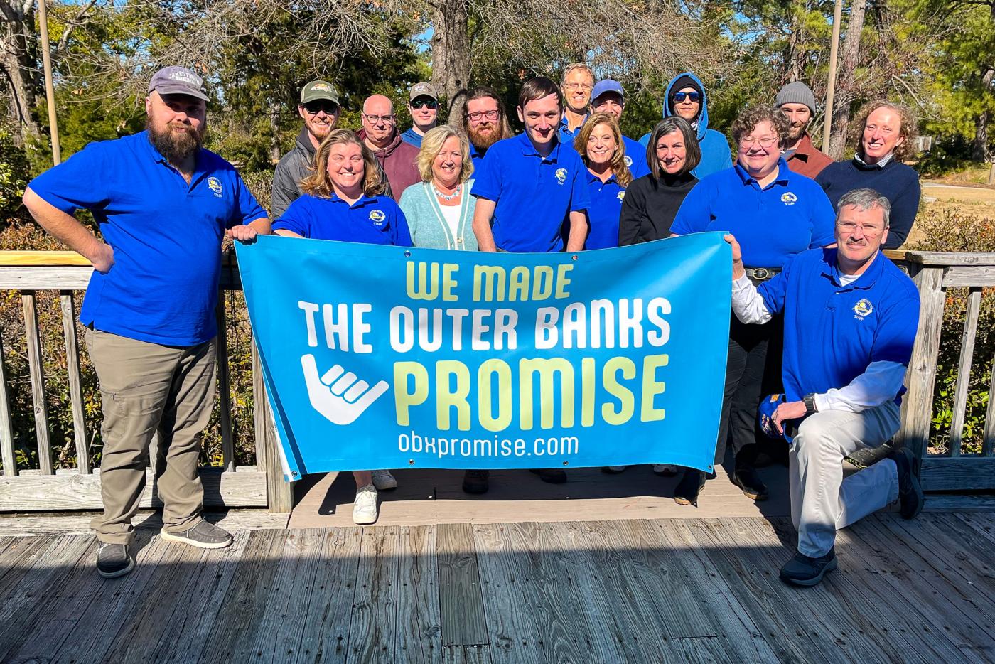 A cheerful group in blue shirts stands on a sunny wooden deck, holding an “Outer Banks Promise” banner with obpromise.com.